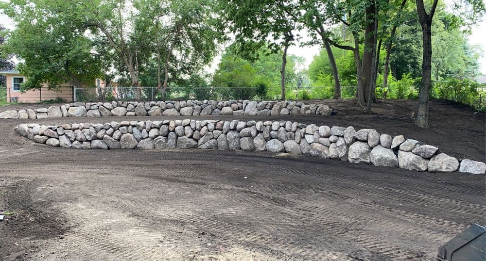 Curved stone retaining wall in a landscaped garden with freshly turned soil and trees.