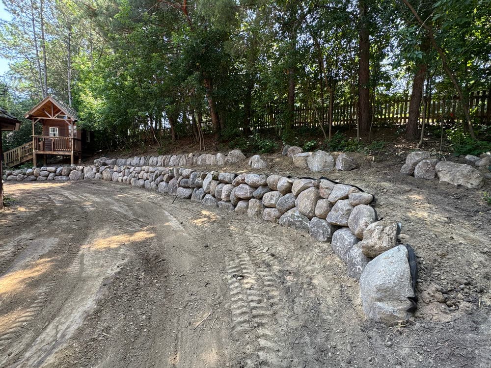 Landscape with a stone wall beside a dirt path and trees, near a wooden cabin.
