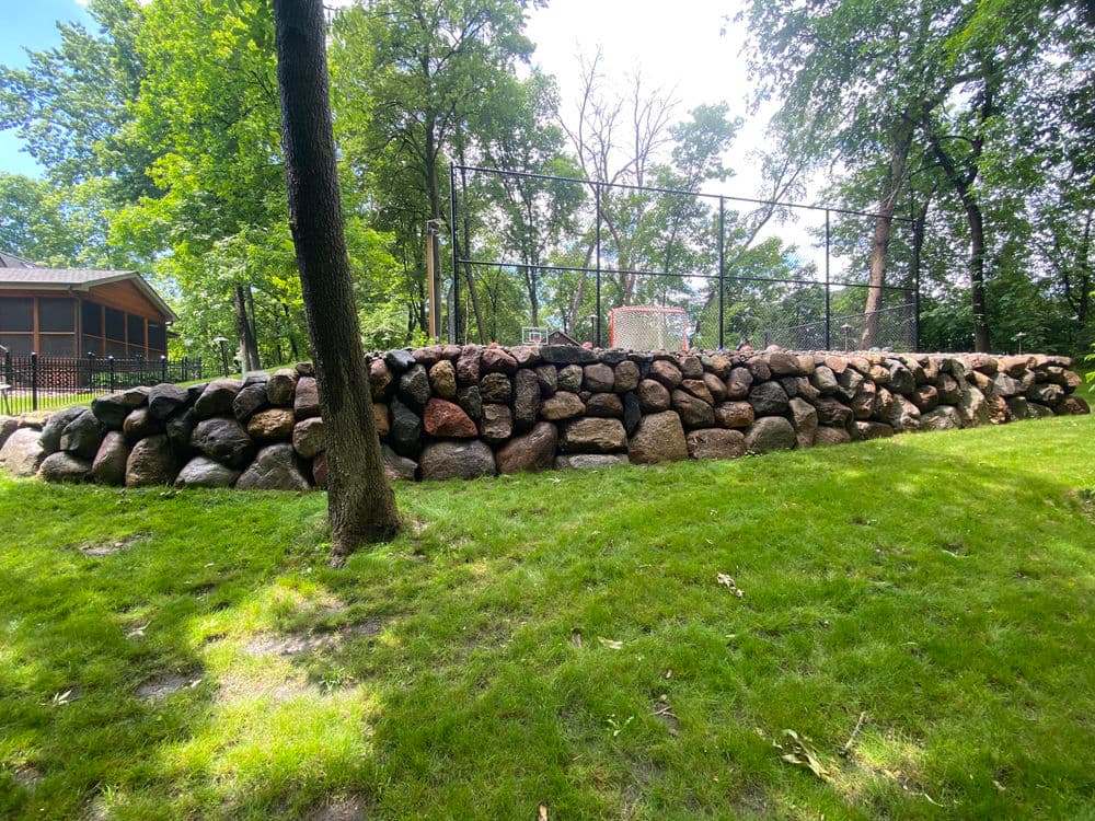 Stone wall surrounding a grassy area near a basketball court in a wooded park.