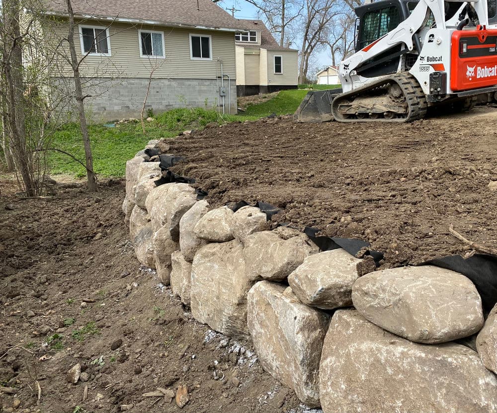 Bobcat excavator on a dirt landscape with stone retaining wall under construction.