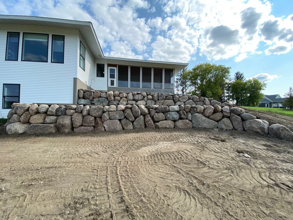Landscape featuring a stone retaining wall beside a modern home with a clear sky.