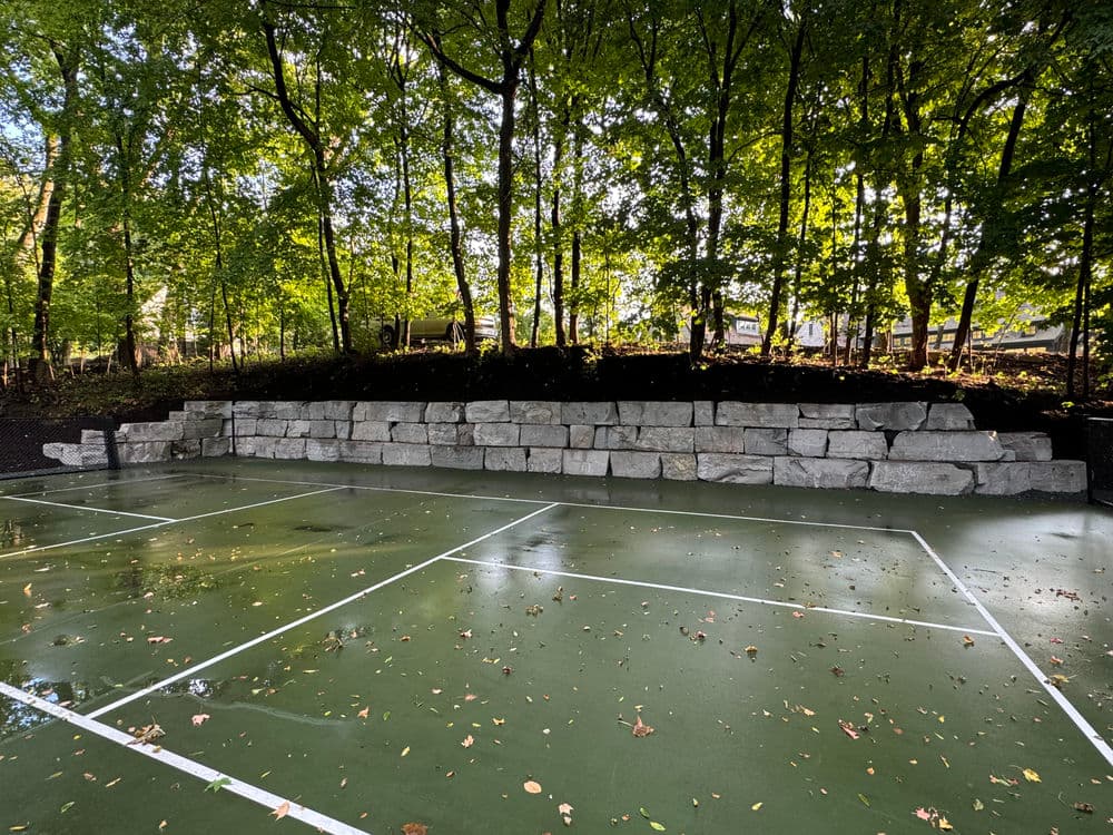 Stone retaining wall behind a green tennis court surrounded by trees.