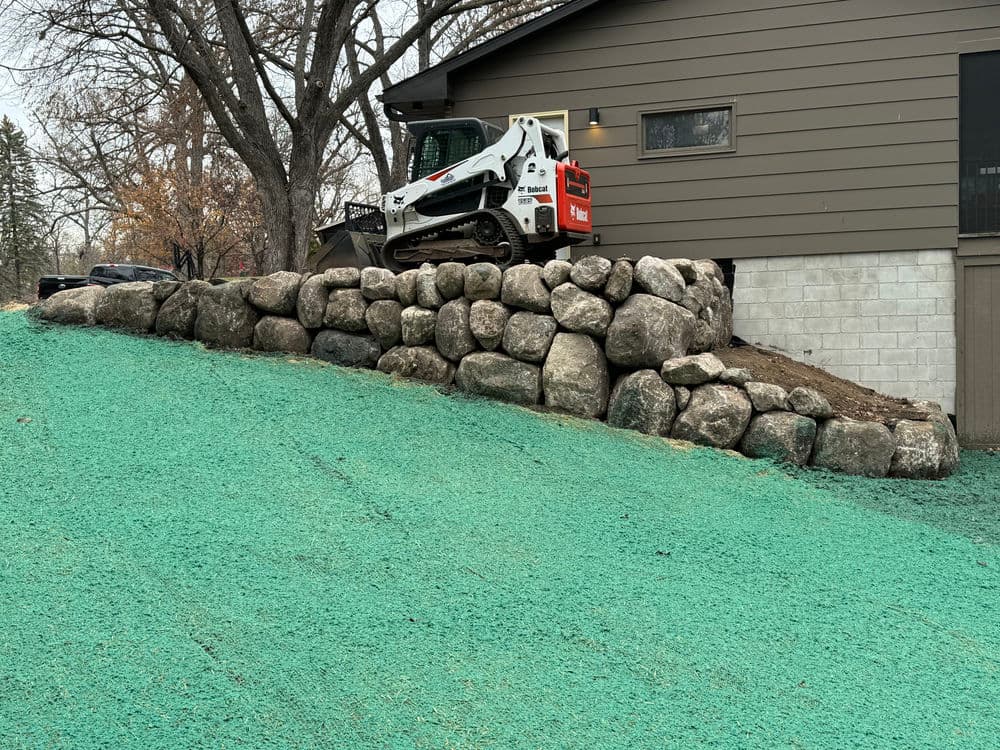 Skid steer on rocky slope near a modern brown house with green turf.