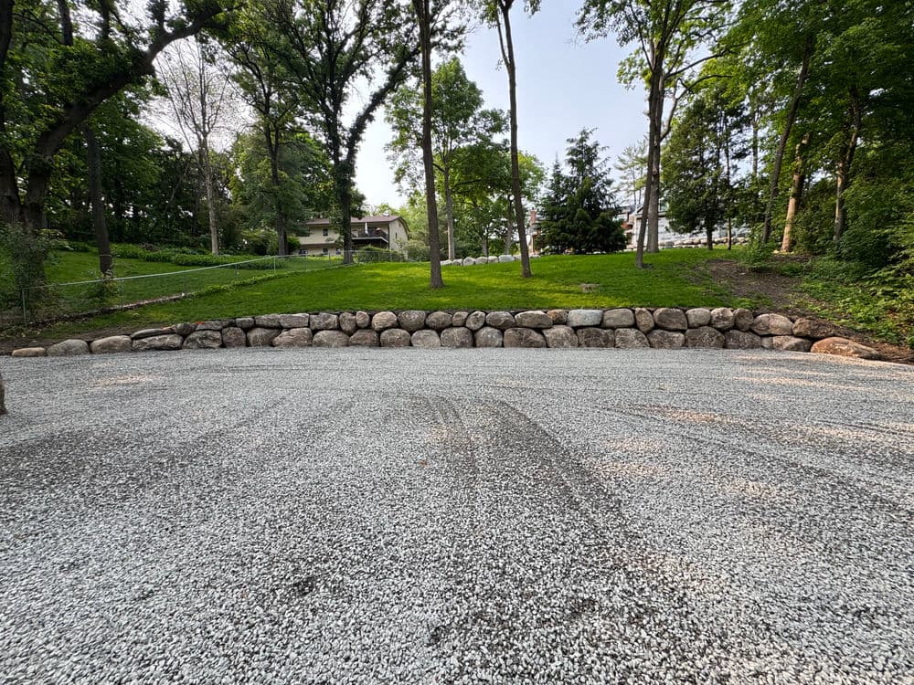 Gravel area with a sturdy stone wall, surrounded by trees and a grassy backdrop.