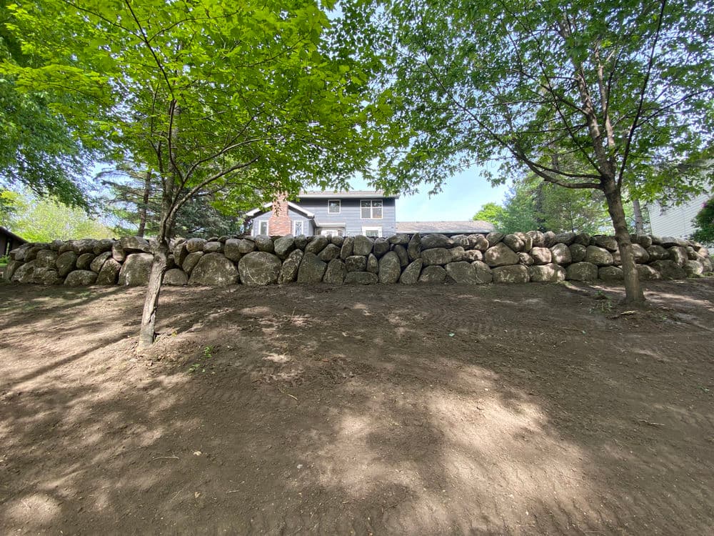 Natural stone retaining wall with trees and a house in the background on a sunny day.