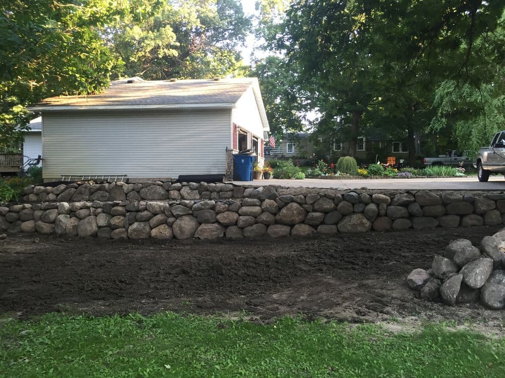 Home exterior with a stone wall, freshly leveled ground, and landscaped garden in the background.