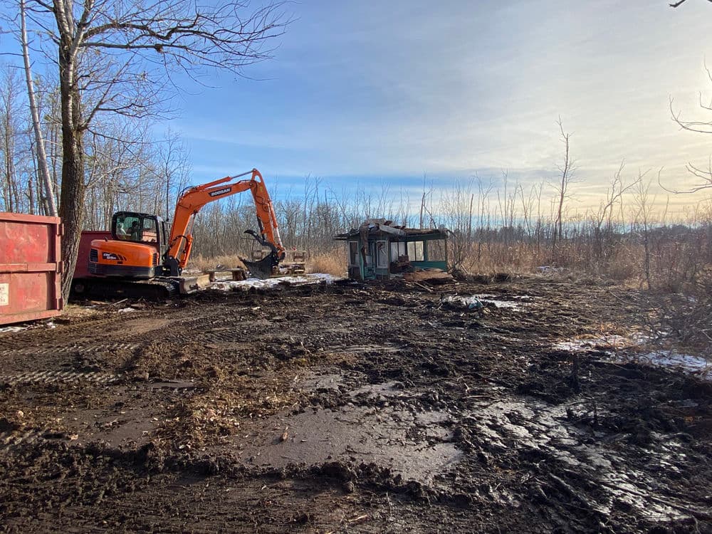 Excavator clearing debris in a muddy construction site with a dilapidated building.