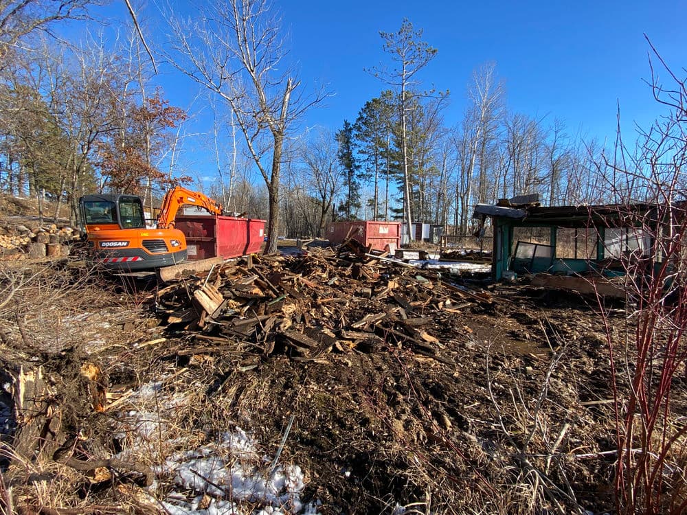 Excavator and debris piles at a construction site in a wooded area during winter.
