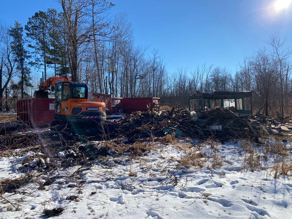 Heavy machinery clearing debris in a snowy forest area with wooden piles and dumpsters.