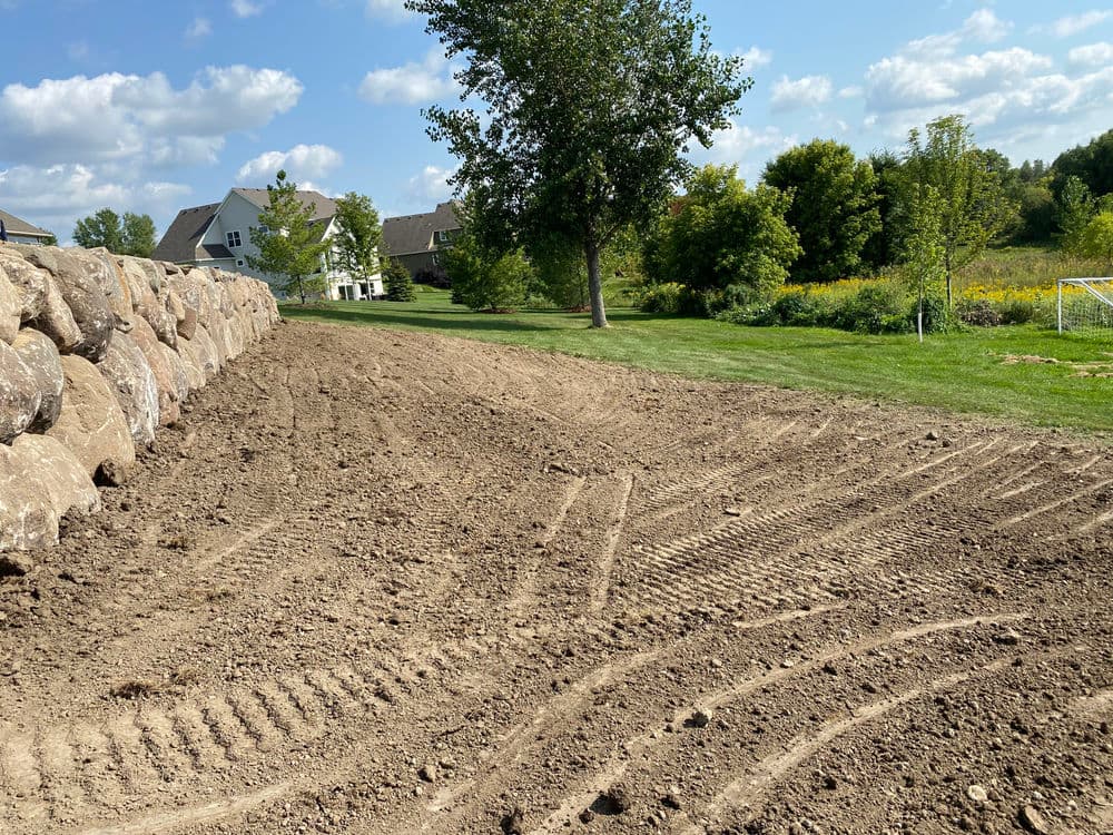 Freshly tilled earth beside a stone wall, with trees and a house in the background.