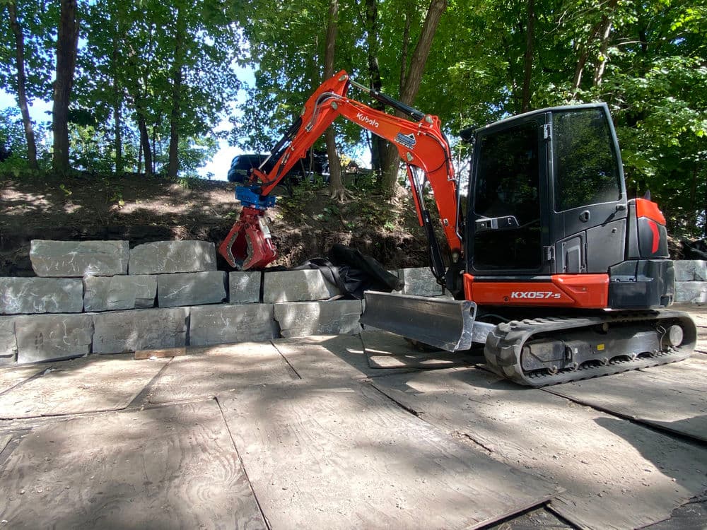 Excavator lifting stone blocks in a wooded area, showcasing construction and landscaping work.