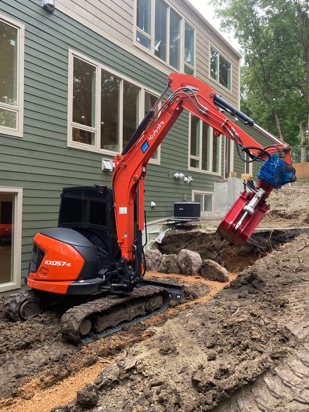 Compact excavator using a hydraulic attachment, digging near a modern house.