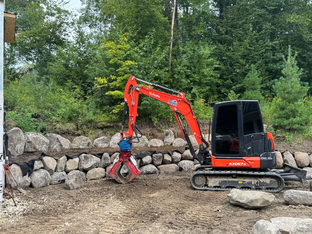 Excavator working on landscaping project with large rocks in forested area.