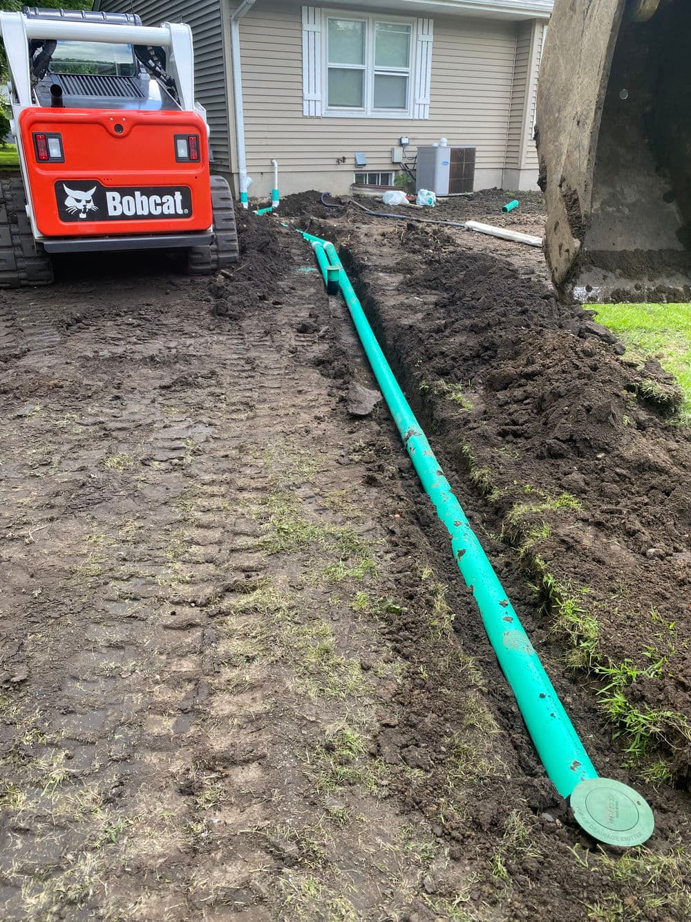 Bobcat excavator next to a trench with green PVC pipes for utility installation.