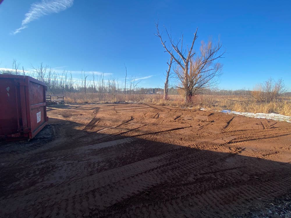 Empty construction site with dirt ground, a red dumpster, and a bare tree under blue sky.