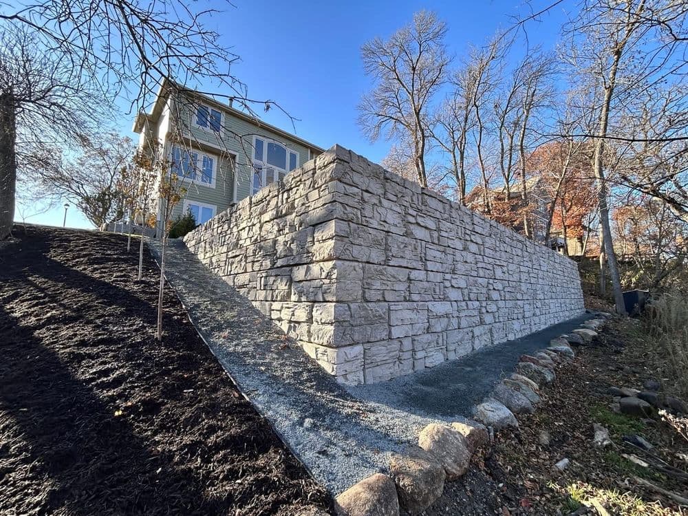 Retaining wall made of stone beside a house, surrounded by trees and landscaping.